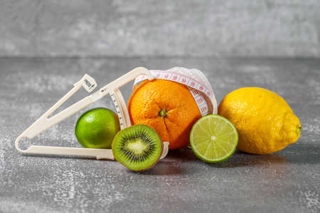 An orange wrapped with a measuring tape and a caliper surrounded by fresh fruits on a gray concrete background. The concept of slimming, remove cellulite, bring the figure into shape.の写真素材