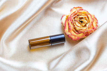 Container for storing cosmetics made of dark glass lie on the folds of silk satin fabric, surrounded by dried rose flowers. Scent fragrance cosmetic beauty product.の写真素材