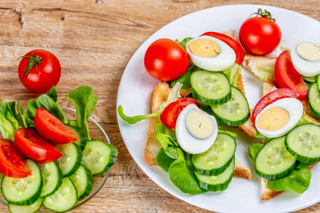 Fried white bread toasts with vegetable and egg slices and cucumber and tomato salad on a plate on a wooden backgroundの写真素材