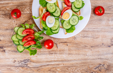 Fried white bread toasts with vegetable and egg slices and cucumber and tomato salad on a plate on a wooden background. Top view.の写真素材