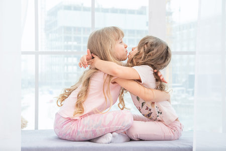 Cute little siblings sitting on windowsill at home hugging, portrait of pretty little girls on room sill in winter. Adorable pensive child in pink.の写真素材