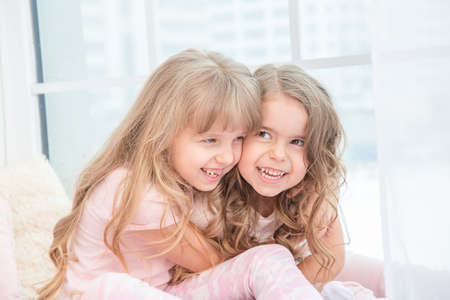 Cute little siblings sitting on windowsill at home hugging, portrait of pretty little girls on room sill in winter. Adorable pensive child in pink.の写真素材