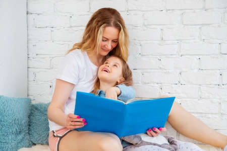 Happy Caucasian mom reads a cheerful fairy tale in a book to her daughter and smiles while sitting...