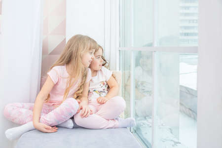 Cute little siblings sitting on windowsill at home looks out window to snow, portrait of pretty little girls on room sill in winter. Adorable pensive child in pink, cute toddler during contemplation.の写真素材