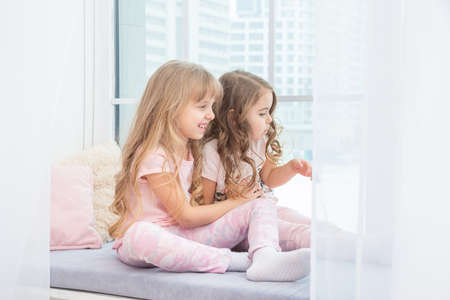 Cute little siblings sitting on windowsill at home looks out window to snow, portrait of pretty little girls on room sill in winter. Adorable pensive child in pink, cute toddler during contemplation.の写真素材
