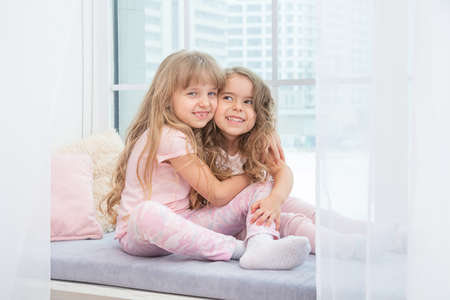 Cute little siblings sitting on windowsill at home hugging, portrait of pretty little girls on room sill in winter. Adorable pensive child in pink.の写真素材