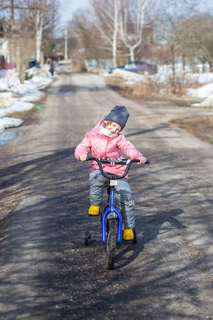 Little girl learns to ride bicycle with safety wheels on the road in the village when all the snow has not yet meltedの写真素材