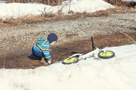 Children balance bike lies on a snowdrift in early spring, a little boy in a spring overalls is playing nearbyの写真素材