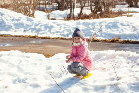 Little caucasian girl 5 years old in a pink coat plays in the snow on her plot of a country house in the springの写真素材