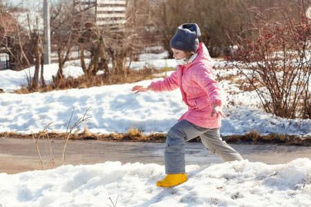 Little caucasian girl 5 years old in a pink coat plays in the snow on her plot of a country house in the springの写真素材