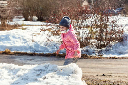 Little caucasian girl 5 years old in a pink coat plays in the snow on her plot of a country house in the springの写真素材