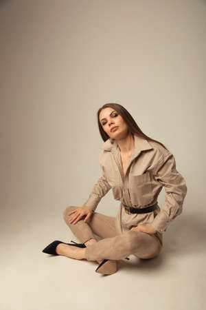 Portrait of young caucasian brunette woman in a beige blouse posing on the floor and looking at the camera over beige backgroundの写真素材
