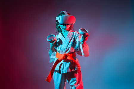Studio Portrait of young caucasian girl in karate uniform playing video games with virtual reality headset or practicing online with a trainer on blue and red background. Future technology concept. VRの写真素材