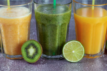 Three glasses in a row with orange juice and banana orange kiwi and spinach smoothie surrounded by halves of fruit on a gray concrete background. The concept of losing weight and proper nutrition.の写真素材