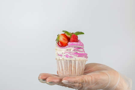 A female hand in transparent vinyl gloves holds a cupcake with Strawberries and pink cream. Close-up...