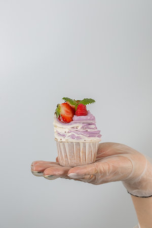 A female hand in transparent vinyl gloves holds a cupcake with Strawberries and pink cream. Close-up white background.の写真素材