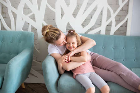 Little caucasian girl sitting together with mother on sitting on the sofa in living room, hugging and kissing each other. Spending time with children at home.の写真素材