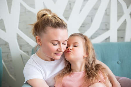 Little caucasian girl sitting together with mother on sitting on the sofa in living room, hugging...
