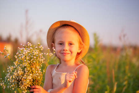 happy little caucasian girl 5 years old with a bouquet of flowers of chamomiles posing in the meadow looking at the camera, a portrait at sunsetの写真素材