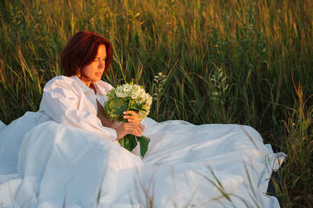 Charming caucasian woman 30-35 years old in white bed in a field with a bouquet of hydrangea in a white men's shirt at sunset in summer, beauty in nature concept.の写真素材