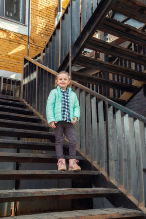 Little caucasian blonde girl in a fashionable youth warm spring outerwear stands on the iron vintage staircase looking at the camera. Pretty beautiful girl in stylish clothes.の写真素材