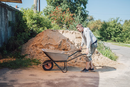 A 30-year-old caucasian man pours sand from a large pile of sand onto a garden wheelbarrowの写真素材