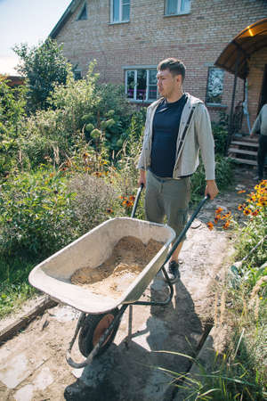 A 30-year-old caucasian man carries a garden wheelbarrow for loading sand to fill the paths with...