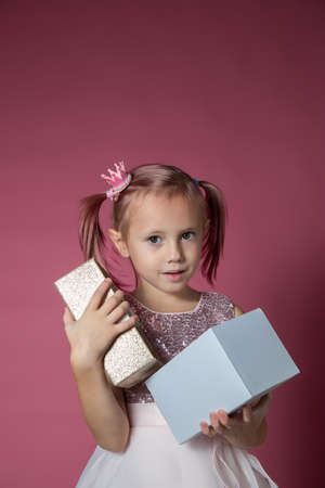 Little caucasian girl in a festive dress with sequins posing opening gift box on a pink background looking at the cameraの写真素材