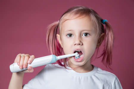 Smiling caucasian little girl cleaning his teeth with electric sonic toothbrush on pink background.の写真素材