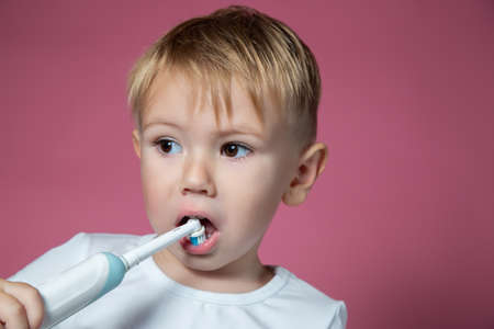 Smiling caucasian little boy cleaning his teeth with electric sonic toothbrush on pink background.