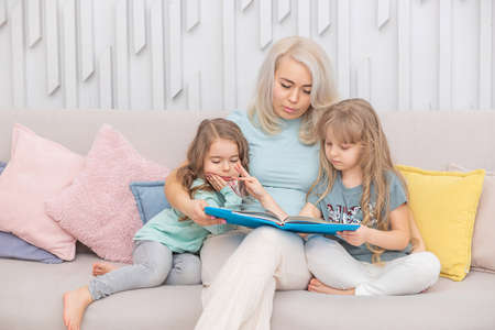 Happy blond mother reading a book to her cute daughters sitting on the sofa in the living room....