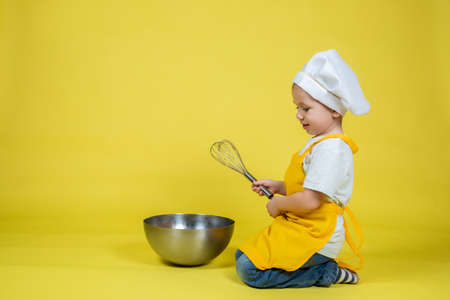 Little caucasian Boy playing chef, boy in apron and chef's hat sitting on the floor with a bowl and...