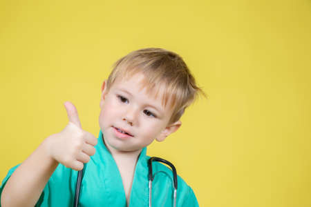 Portrait of cute little caucasian boy dressed in doctors green showing thumb up gesture on yellow backgroundの写真素材