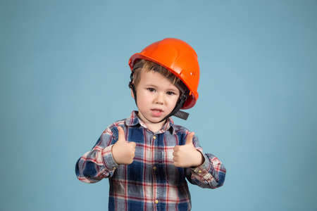 Portrait of adorable little child in a protective orange helmet over blue background showing thumb...
