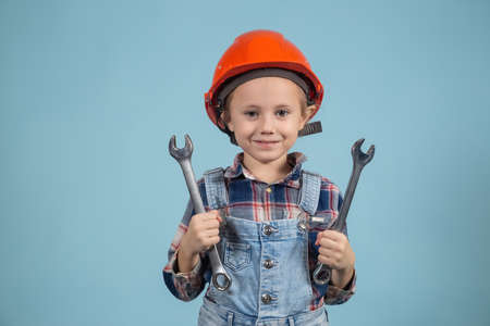 A cute girl in an orange hard hat posing on blue background, holding wrenches in both hands....