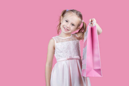 Cute little caucasian girl in a party dress on pink background happy enjoying shopping holding colorful bagsの写真素材