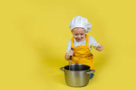 Little caucasian Boy playing chef, boy in apron and chef's hat sitting on the floor with a saucepan and ladle on yellow backgroundの写真素材