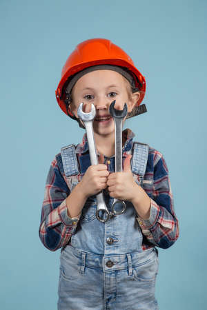 A cute girl in an orange hard hat posing funny on blue background, holding wrenches in both hands. Construction and repair concept.の写真素材