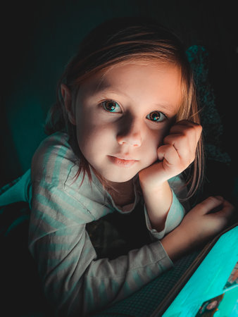 Little caucasian girl looking at a tablet in the evening in the dark on the bed,lifestyle toned photography at homeの写真素材