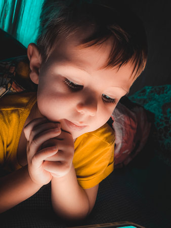 Little caucasian boy looking at a tablet in the evening in the dark on the bed,lifestyle toned photography at homeの写真素材