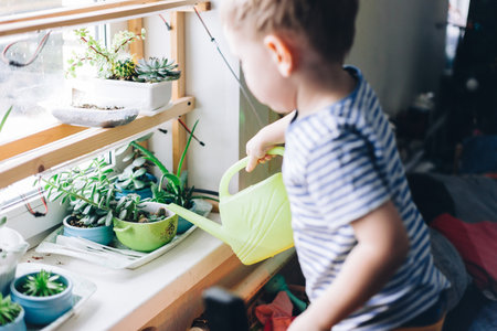 Caucasian boy watering from a yellow watering can home plants on the windowsill, toned lifestyle photography at homeの写真素材