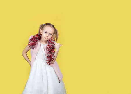 Portrait of cute little child girl in a smart dress and with tinsel making a pointing gesture with place for text over yellow background. The concept of New Year and Christmas.の写真素材