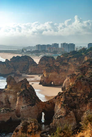 As the sun goes down natural rock archs and cliffs form a scenic view of the beach, a city is visible in the backgroundの写真素材