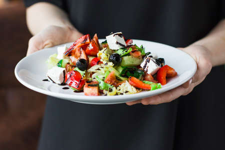 The young female waiter holding in her hands the plate with the Greek saladの写真素材