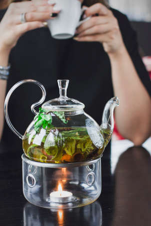 The glass teapot with herbal tea on stand with candle on the background of a young woman with mug of teaの写真素材