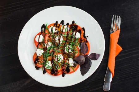 Caprese salad on a white plate with the devices in an orange napkin on the black table. The view from topの写真素材