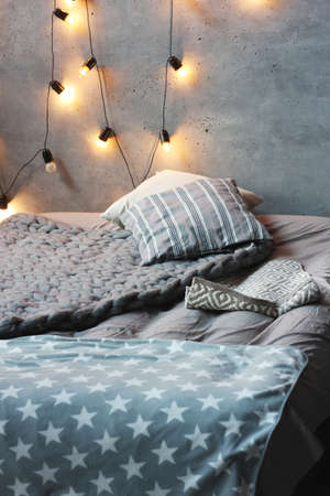 Bed with pillows in loft room, concrete walls with garland of light bulbs. Scandinavian interior