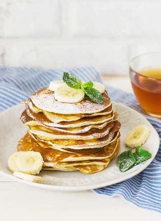 fresh baked pancakes with banana in white plate and cup of tea on blue linen on white backgroundの写真素材