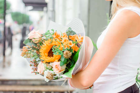 Young woman with blonde hair holding bouquet of flowers rose sunflower on the streetの写真素材