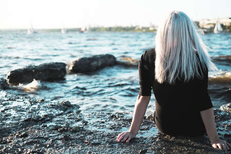 The girl with blond hair sits back on shore of lake sea and looks into the distance at boatsの写真素材